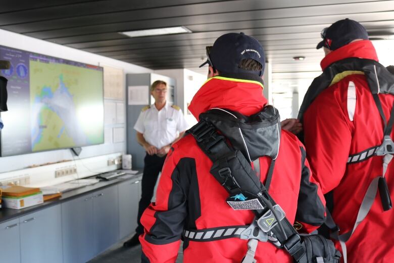 Two pilots in red flotation jackets listen to a cruise ship captain as he stands in front of  map, explaining the journey to come.