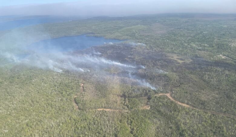 An aerial shot shows blackened portions of forest near a lake, with smoke rising from several areas.