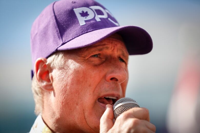 A closeup image shows a man speaking into a microphone and wearing a purple-coloured baseball hat.