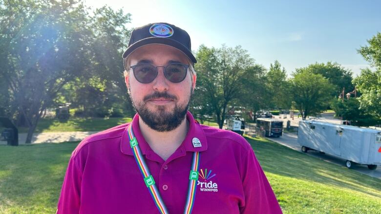 A person with a baseball cap, sunglasses and a beard poses outside under bright sun.