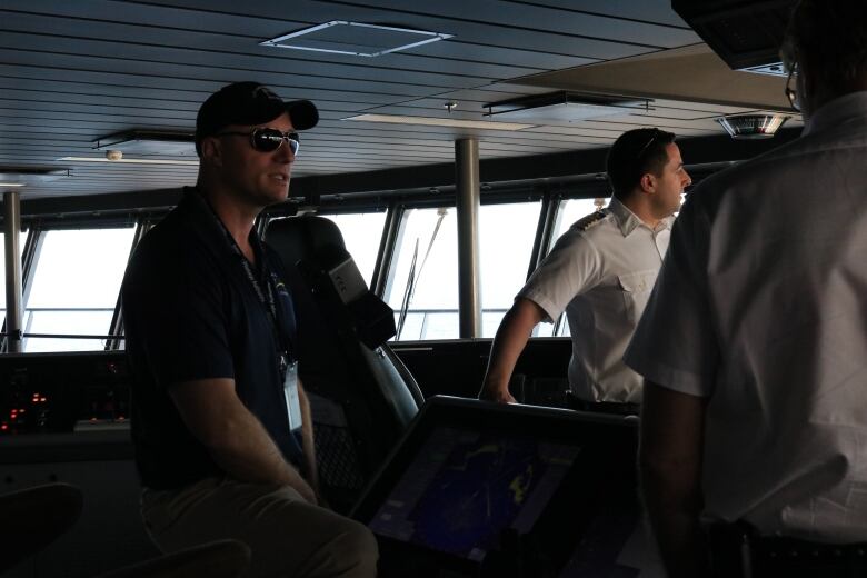 A fit man in sunglasses and a ball cap sits on the bridge of a ship talking to a uiniformed crew. 