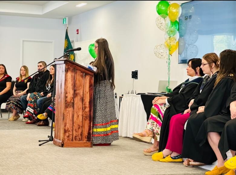 A woman with long brown hair stands at a podium. There are graduates sitting on either side of her.