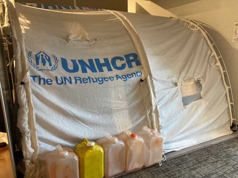 A small white tent with blue lettering. Empty plastic water jugs sit next to the tent.