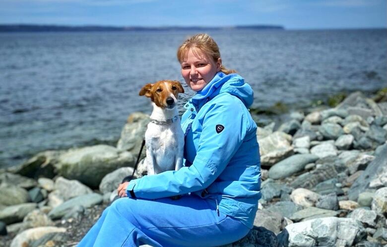 A woman poses for a photograph while sitting on a rocky beach with her dog on her lap.