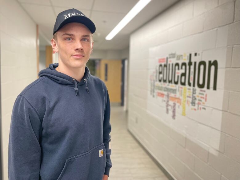 Cruz Pineau, a grade 12 student at Three Oaks Senior High, stands in the hallway at the school. He's wearing a black baseball cap and blue hoodie. 