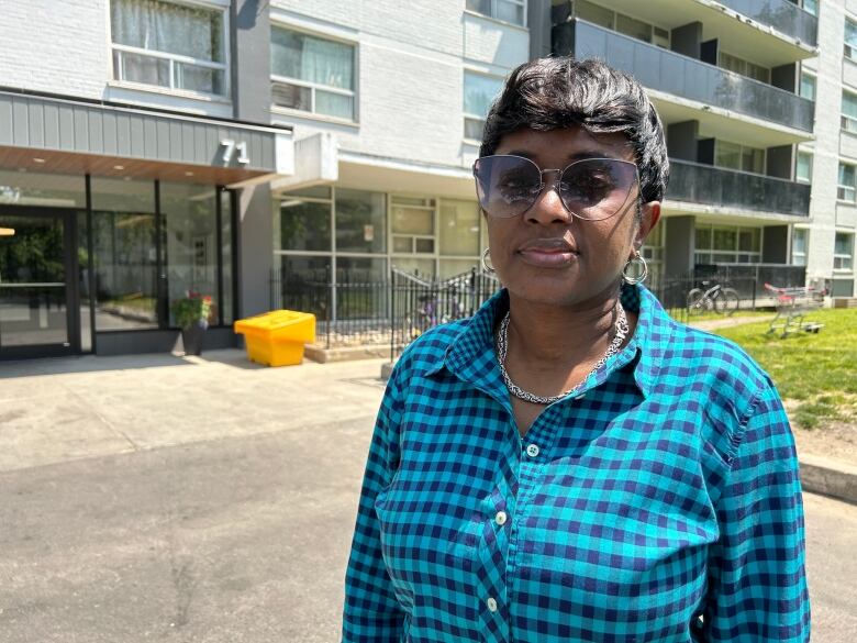 A woman wearing a blue checked shirt and sunglasses stands outside of an apartment building. 