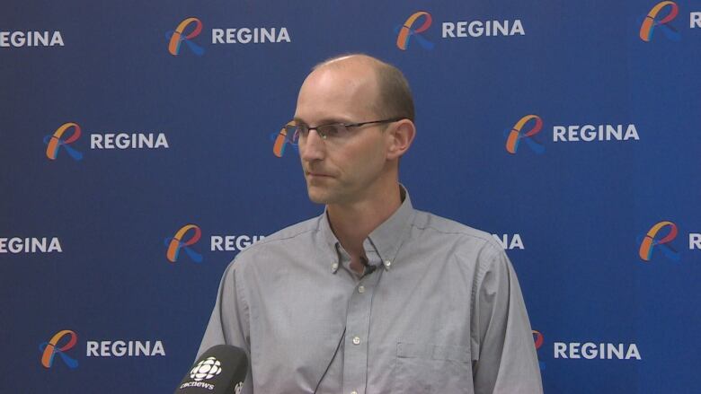 A bald white man with glasses is wearing a grey button-up shirt, with the top button undone. He standing in front of a large blue poster with the word 'Regina' on it multiple times.