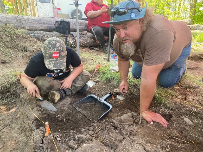 A man and a young boy kneeling in the dirt with tools to do an archaeological dig. 