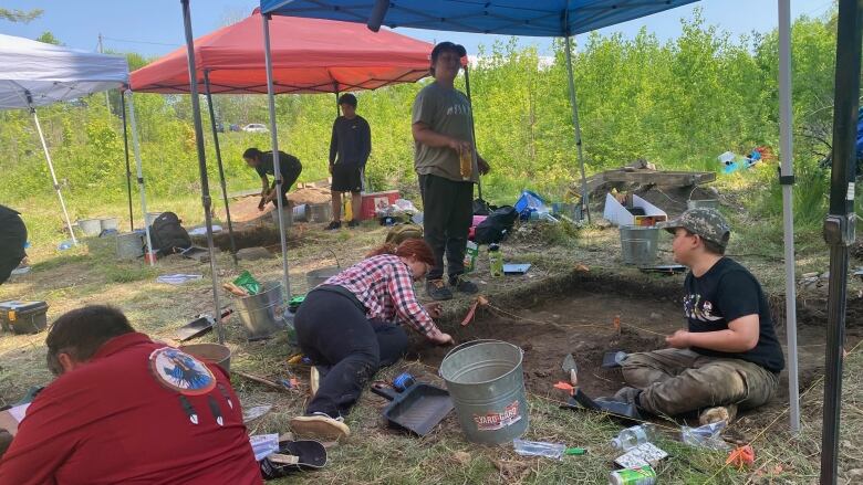 People kneeling and digging in dirt, with tents overhead. 