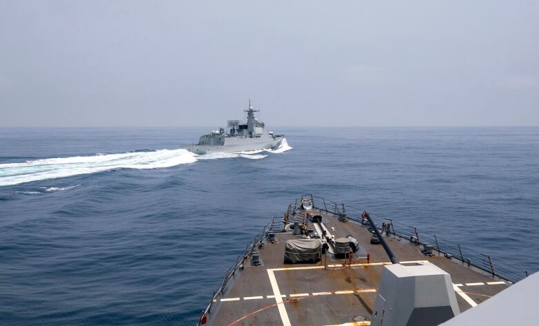 A navy ship cuts in front of the bow of a large ship. A wake trails the boat.