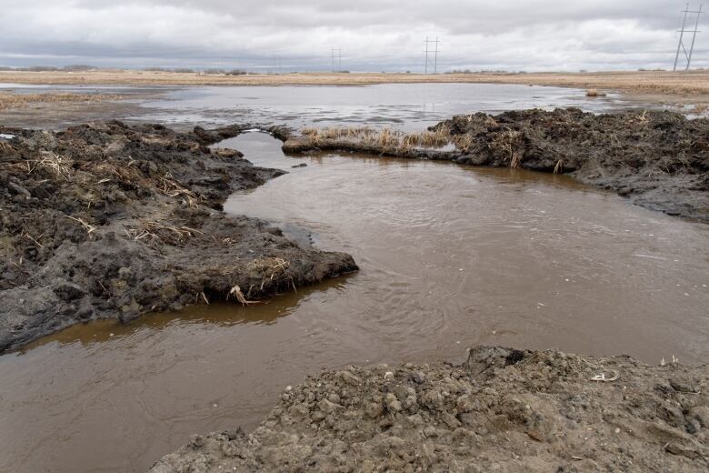 A muddy drainage ditch is filled with water that is flowing into another muddy ditch