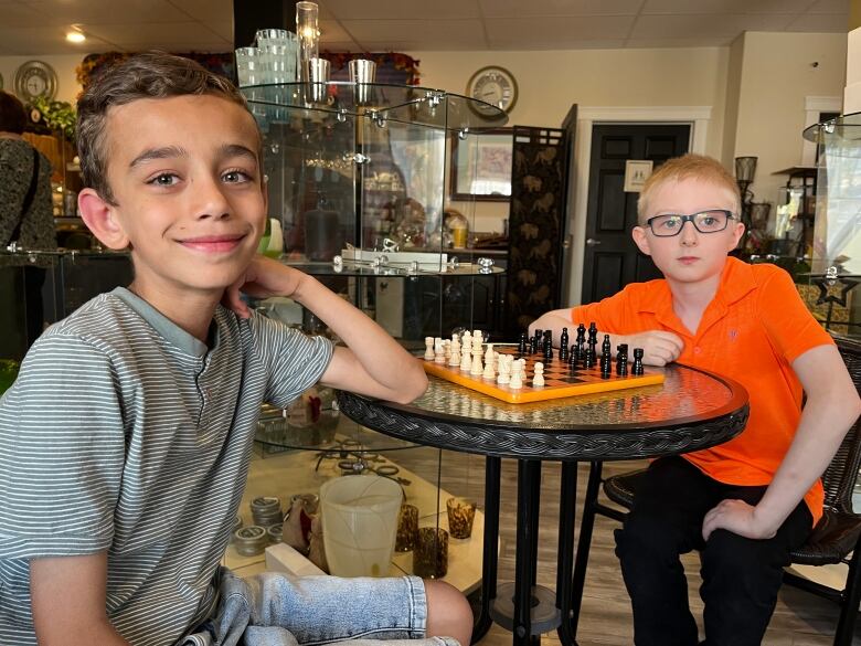 Two boys sit around a table with a chess board. 