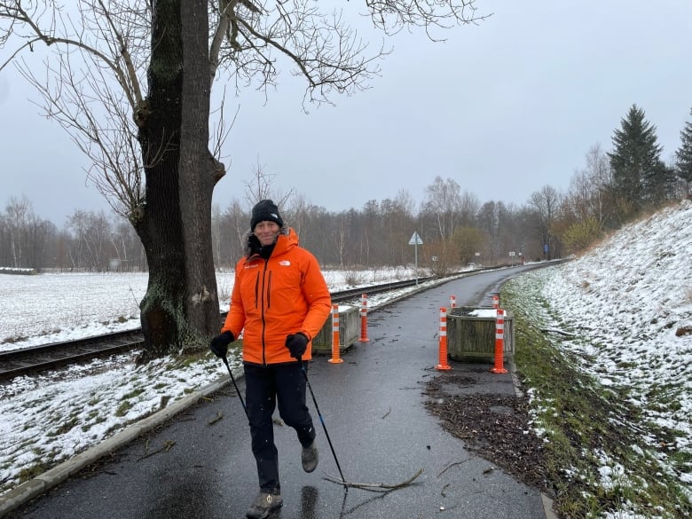 A man in a bright orange rain, black pants and a black toque walks down a cement path next to train tracks and a snowy hill.