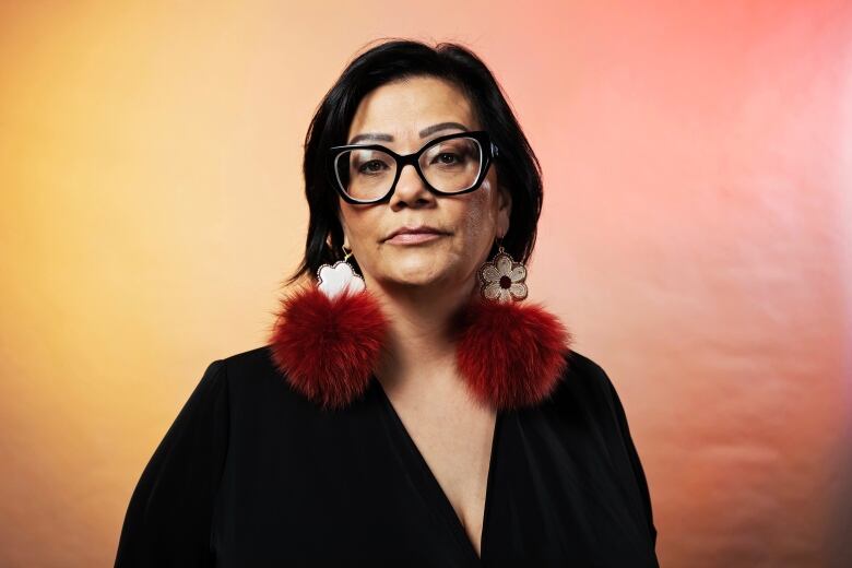 Head and shoulders image of a woman staring straight ahead, with dark hair and wearing dark glasses, a dark blouse and large red and white earrings.