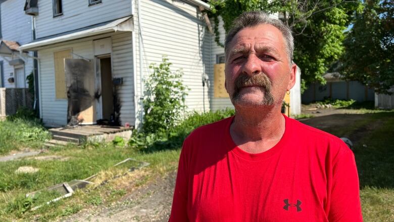 A man in a red shirt is standing in front of a two-storey house. There are boards over the windows, and scorch marks around the door.