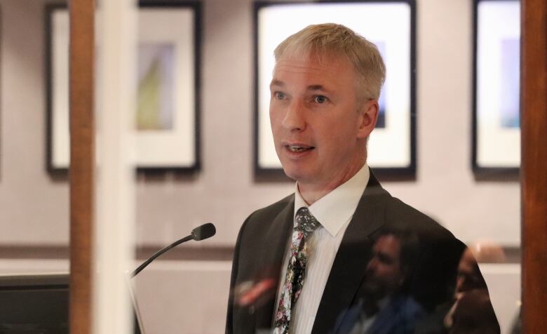 A man speaking at a microphone wearing a suit with framed images on the wall in the background.