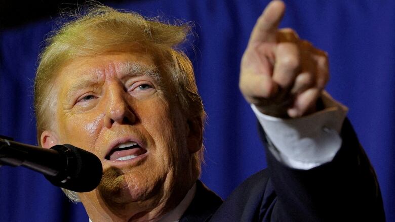 A man with yellow hair and thick face make up gestures as he speaks at a podium.