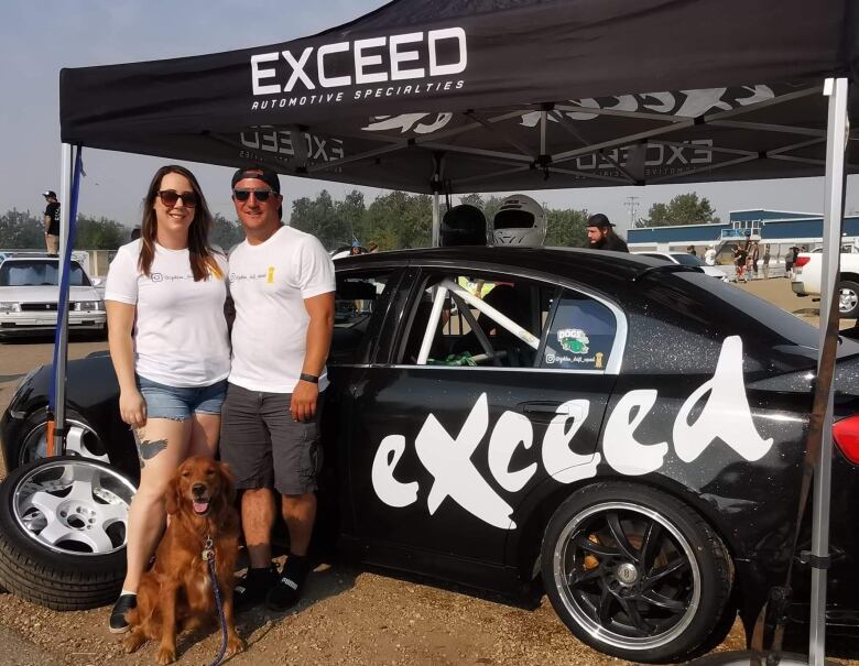 A man stands in front of a black car with a woman and a dog