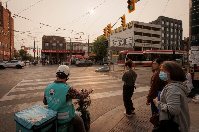 People stand at a crosswalk in Toronto, with grey, hazy skys in the background. 