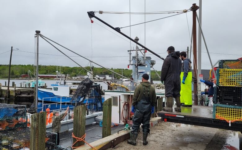 Three fisherman stand on a wharf as another fisherman guides a trap being hauled off a fishing boat.