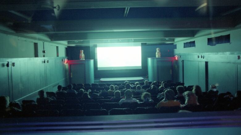 A crowd watches a movie in a theatre. The glare from the screen reflects back towards the audience.