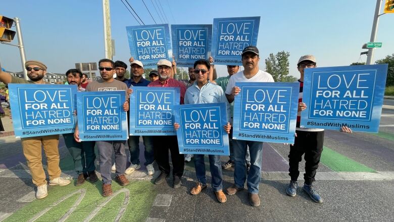 A group of men carrying blue signs reading 'Love for all Hatred for none #StandWithMuslims' stands in the middle of a roadway. 