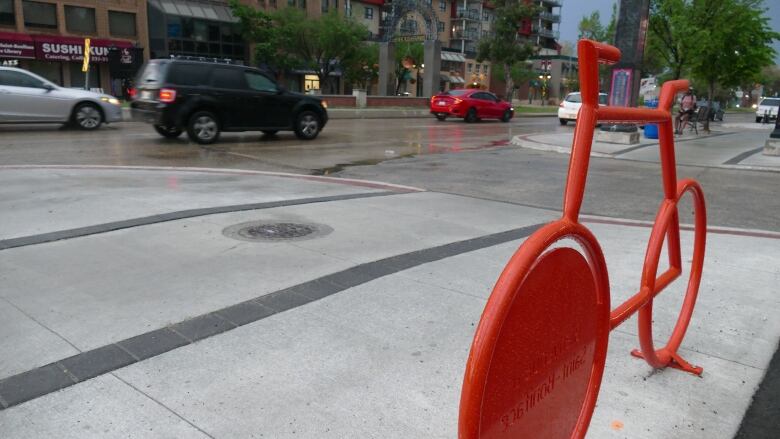 A red metal bicycle rack, in the shape of a bicycle, is seen on the sidewalk in the foreground as traffic moves past in the background.