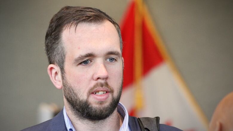 A young man with a beard and moustache, blue striped shirt and blue suit jacket speaks with a red-and-white Canadian maple leaf flag behind him.