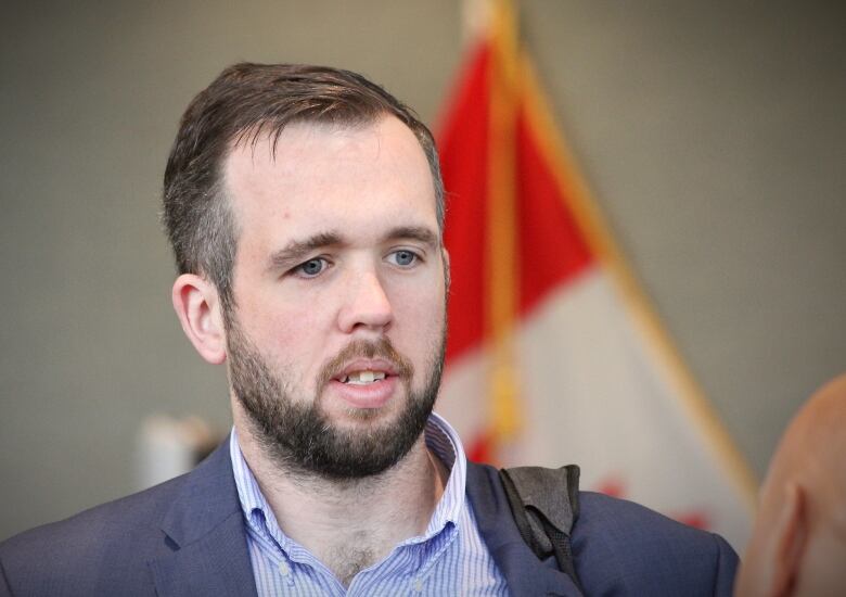 A young man with a beard and moustache, blue striped shirt and blue suit jacket speaks with a red-and-white Canadian maple leaf flag behind him.