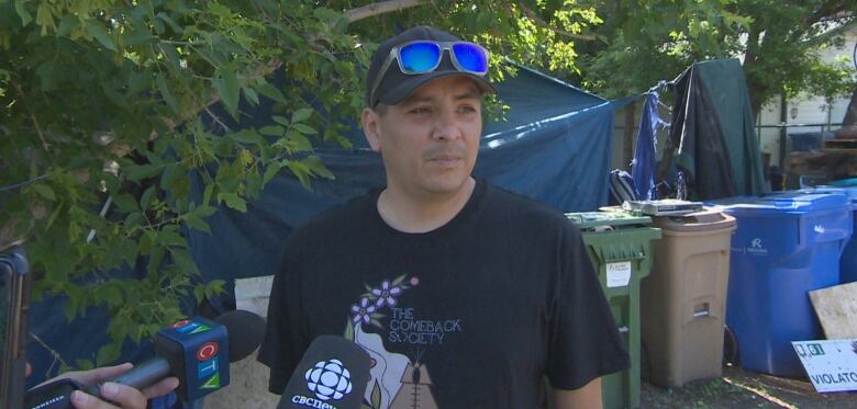 A man in a black shirt with a hat and sunglasses resting on the hat is being interviewed. 