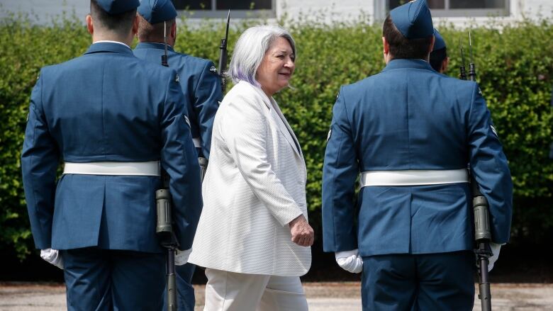 Woman in white suit walks past four people in military garb, with their backs to the camera.