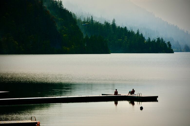 Two people rest on a lakeside quay as they watch a firefighting helicopter take off.