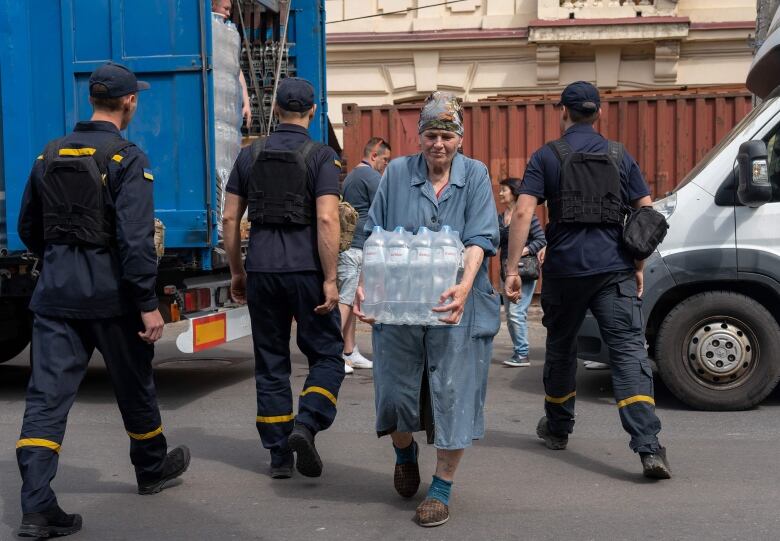 An older woman wearing a headcovering walks backs from a delivery track carrying a case of bottled water as officials in uniform are shown with their backs to the camera.