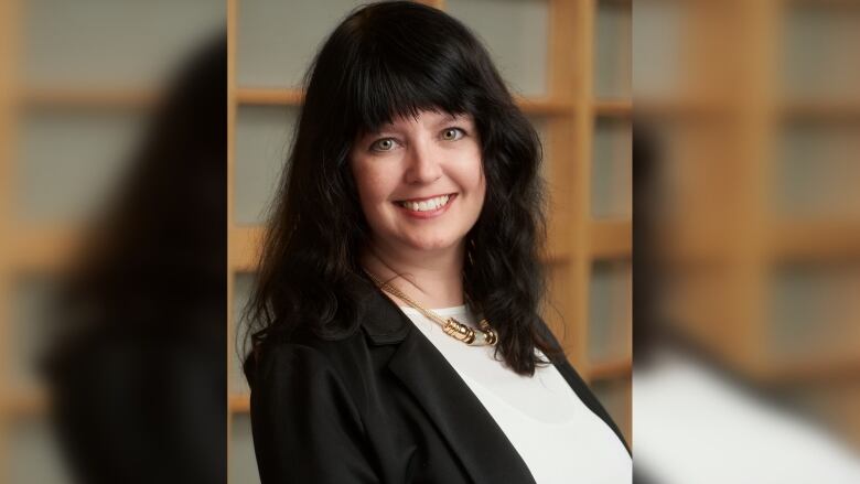 A woman in long black hair and wearing a black blazer over a white blouse smiles.