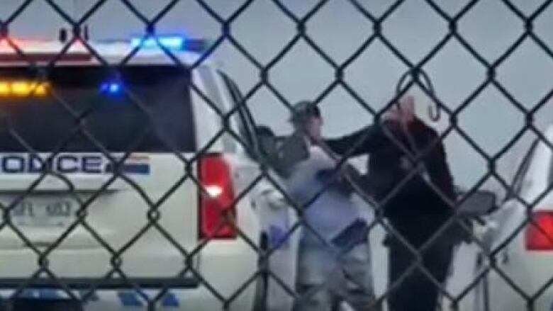 A video taken from behind a chain link fence shows two white RCMP officers arresting a black man. His face and upper body has been pushed up against the car.