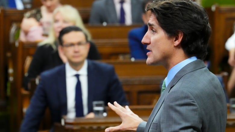 A political leader speaks in a legislature with the opposition leader in the background.