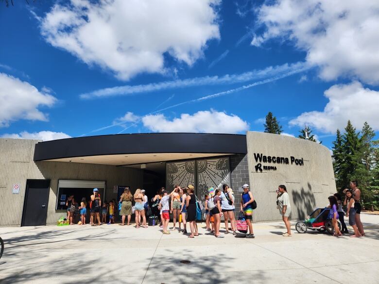 Families, teenagers and children line up outside a concrete building with a sign on the front marking it as Wascana Pool. 