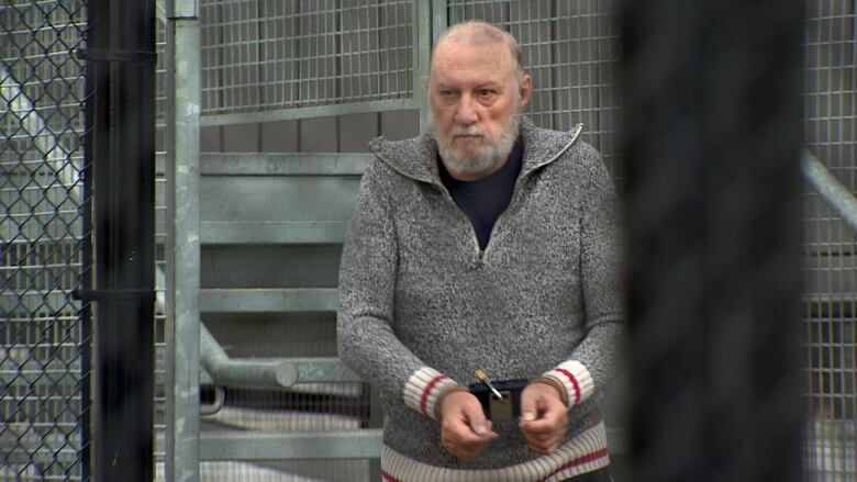 An elderly man in a grey sweater is seen in handcuffs behind a fence outside the courthouse in Iqaluit. 