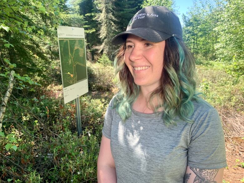 Woman in ball cap and a t-shirt stands in the forest next to a sign on a blue sky day.