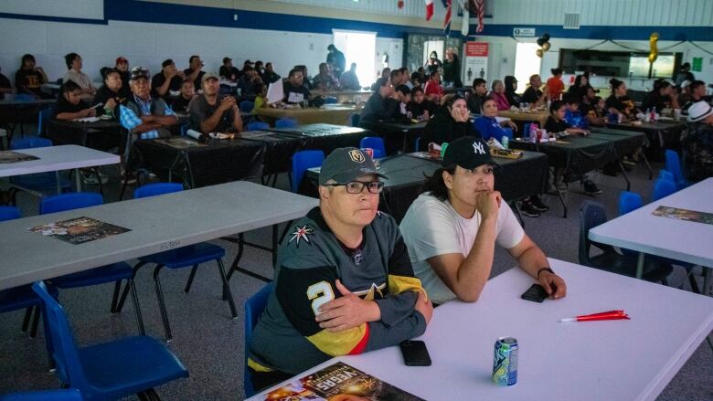 A group of people sit in a large hall wearing Las Vegas Golden Knights gear. 