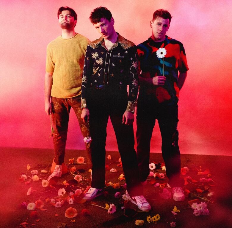 Three men stand among a pile of flowers in a promotional shot of a band.