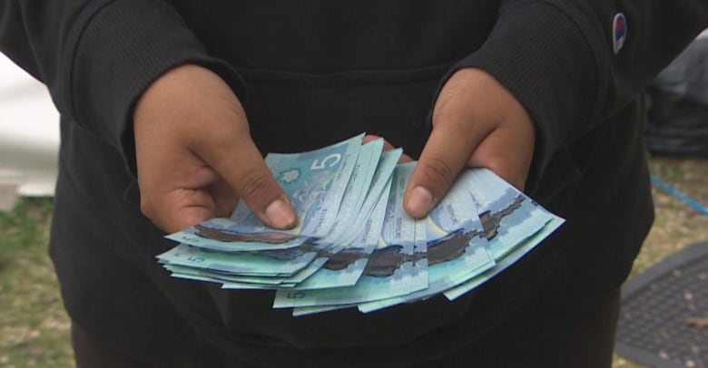 Two hands are pictured showing off a stack of five dollar bills to the camera.