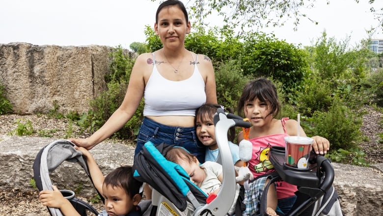 A woman is pictured smiling to the camera, holding a stroller with two kids seated inside. Two older children stand next to her smiling and holding onto the stroller as well.