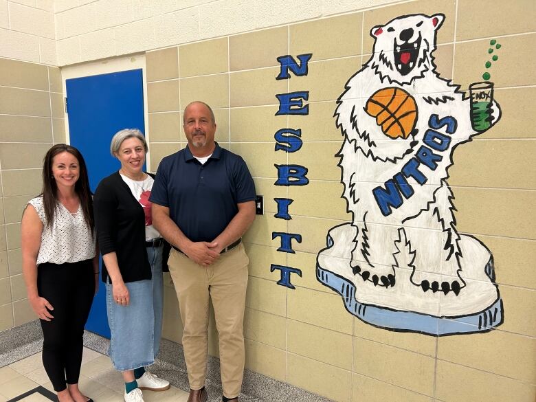 Two women and a man standing inside a school with a painted image of a polar bear on the wall.