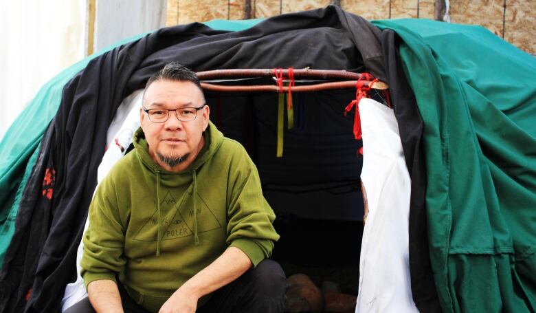 A man in a green sweater sits in front of a large tent. 