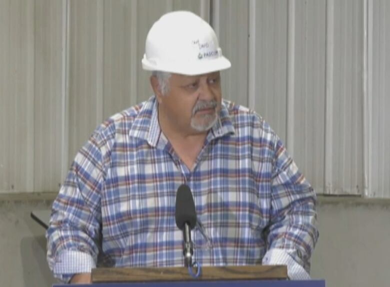 A man in a hard hat and checkered shirt speaks behind a podium and microphone.