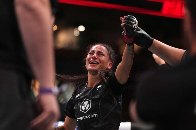 A woman in MMA gear has her hand raised by a trainer as she smiles after winning a fight.