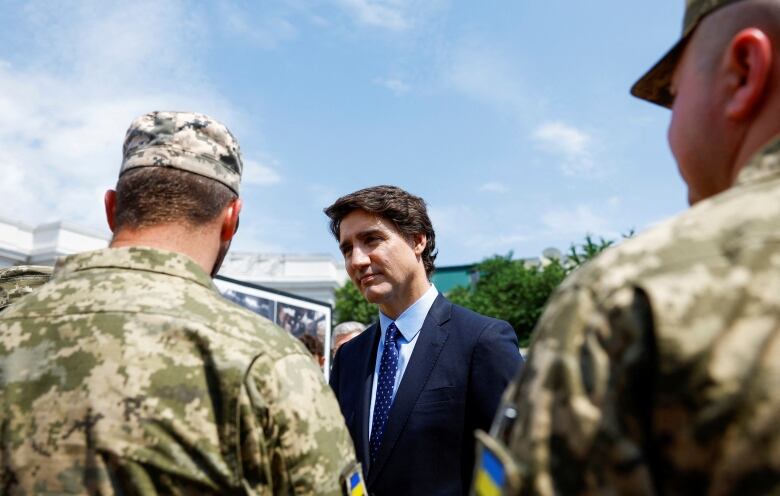 A man with brown hair, wearing a navy suit, speaks with soldiers.