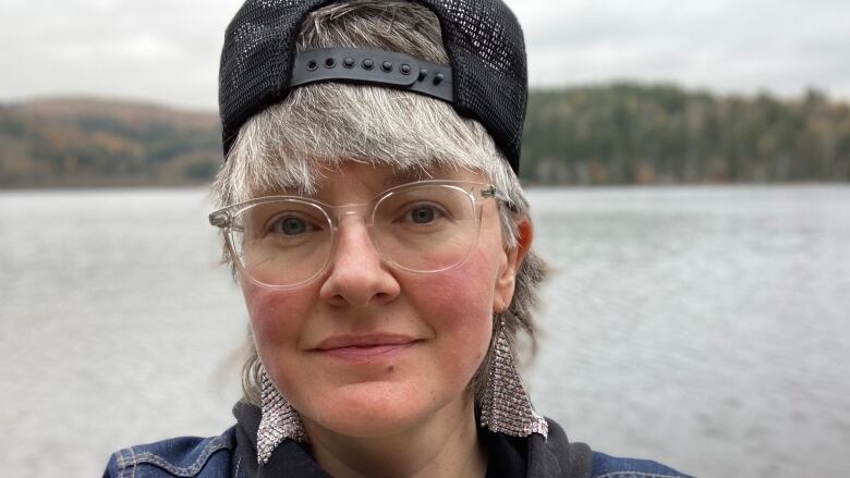 A woman with short hair and glasses and a backwards hat looks at the camera