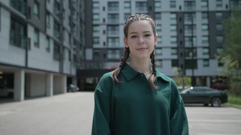 A young woman in a polo shirt with her hair in braids stands in front of an apartment complex.